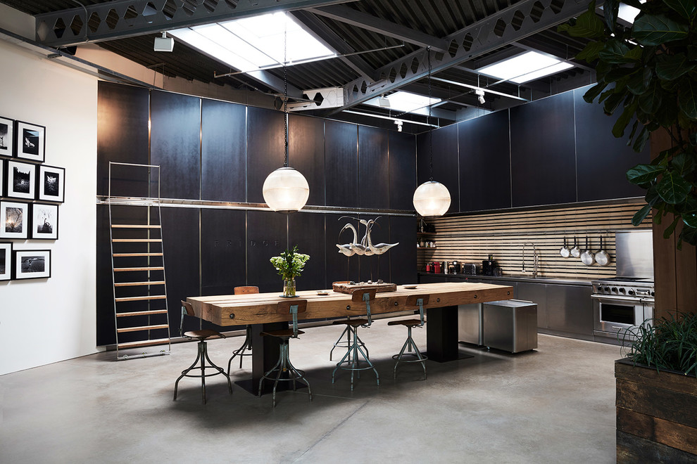 This is an example of an expansive industrial single-wall eat-in kitchen in London with flat-panel cabinets, black cabinets, stainless steel appliances, concrete floors, grey floor, a drop-in sink, stainless steel benchtops and brown splashback.