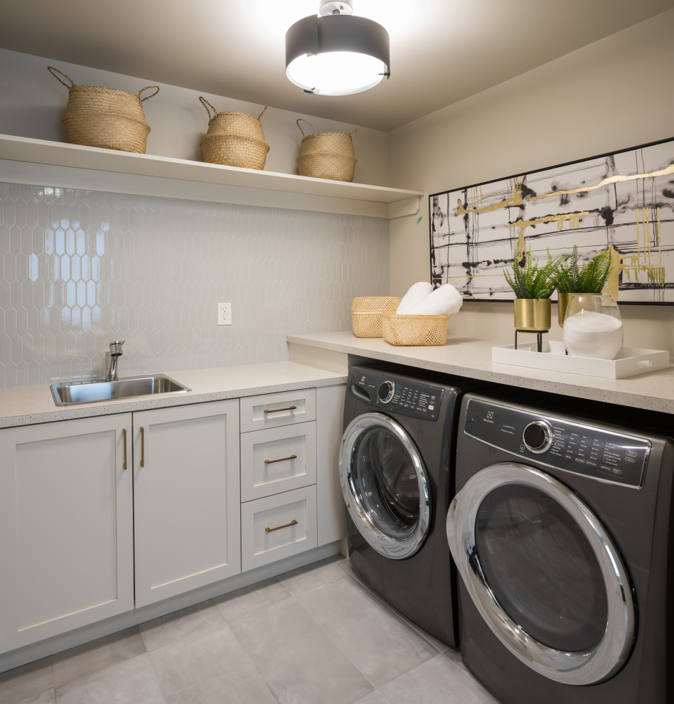 Photo of a large transitional l-shaped dedicated laundry room in Calgary with a single-bowl sink, recessed-panel cabinets, grey cabinets, marble benchtops, grey splashback, ceramic splashback, beige walls, ceramic floors, a side-by-side washer and dryer, grey floor and multi-coloured benchtop.