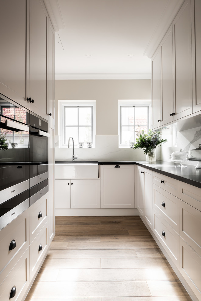 Design ideas for a transitional l-shaped kitchen in Perth with a farmhouse sink, shaker cabinets, white cabinets, white splashback, stone slab splashback, medium hardwood floors, brown floor and black benchtop.