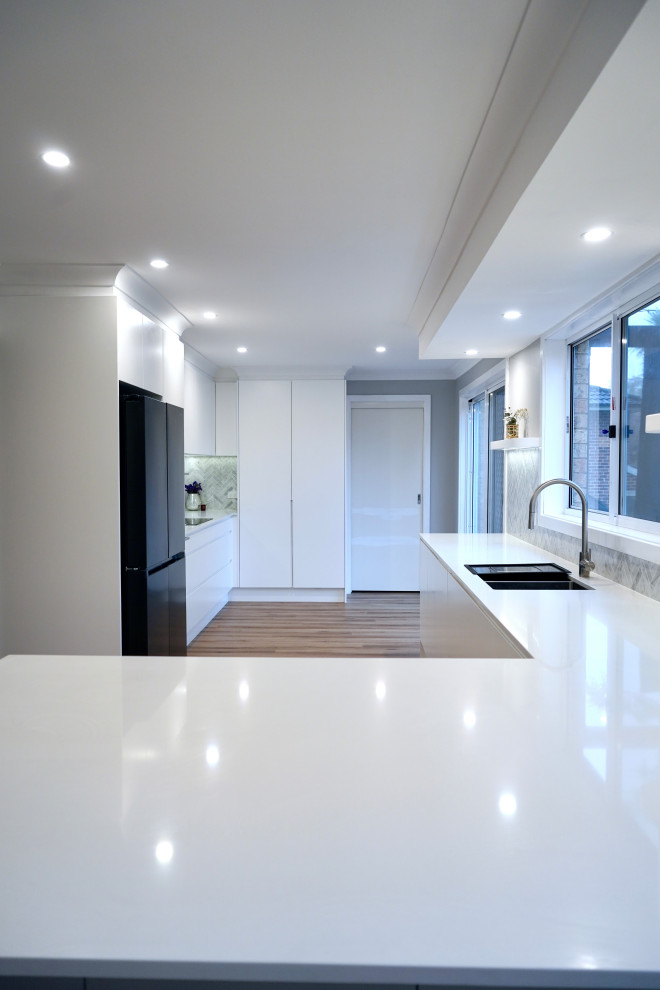 Large modern galley kitchen/diner in Sydney with a built-in sink, flat-panel cabinets, white cabinets, engineered stone countertops, grey splashback, marble splashback, black appliances, light hardwood flooring, brown floors and white worktops.
