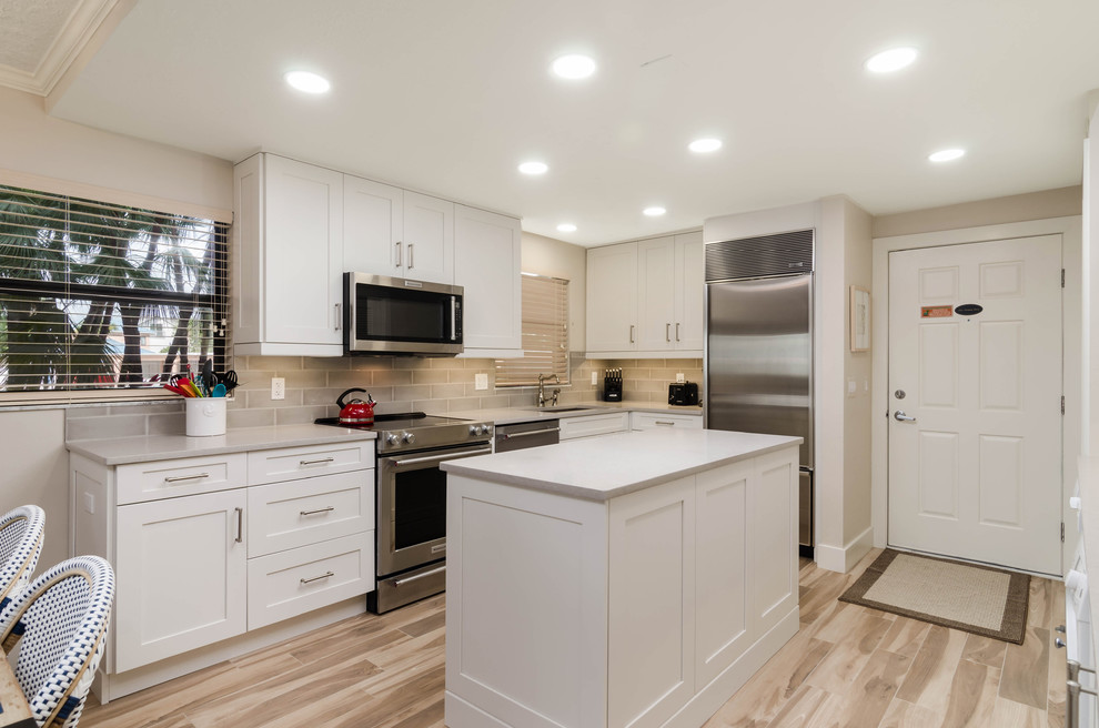 This is an example of a mid-sized beach style l-shaped open plan kitchen in Miami with an undermount sink, shaker cabinets, white cabinets, quartz benchtops, grey splashback, subway tile splashback, stainless steel appliances, laminate floors, with island, beige floor and grey benchtop.