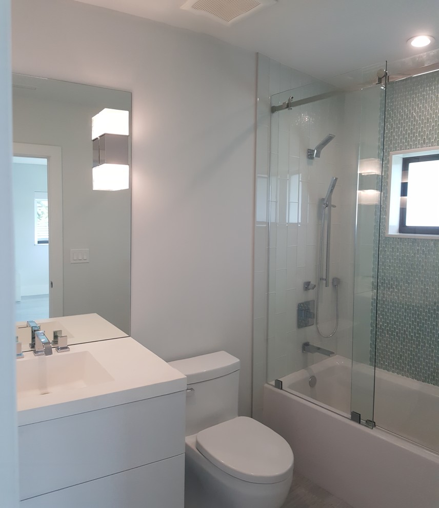 Photo of a mid-sized modern bathroom in Miami with flat-panel cabinets, white cabinets, an alcove tub, a shower/bathtub combo, a one-piece toilet, gray tile, metal tile, grey walls, marble floors, an integrated sink, solid surface benchtops, grey floor and a sliding shower screen.