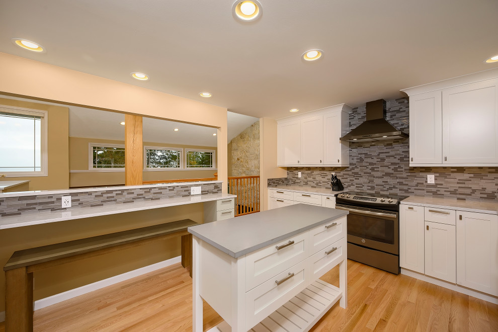 Enclosed kitchen - mid-sized transitional l-shaped medium tone wood floor and brown floor enclosed kitchen idea in Seattle with a farmhouse sink, shaker cabinets, white cabinets, quartz countertops, multicolored backsplash, glass tile backsplash, black appliances, an island and white countertops
