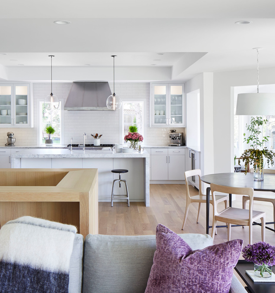 This is an example of a mid-sized scandinavian u-shaped open plan kitchen in Minneapolis with a farmhouse sink, flat-panel cabinets, white cabinets, marble benchtops, white splashback, subway tile splashback, stainless steel appliances, light hardwood floors, with island and beige floor.