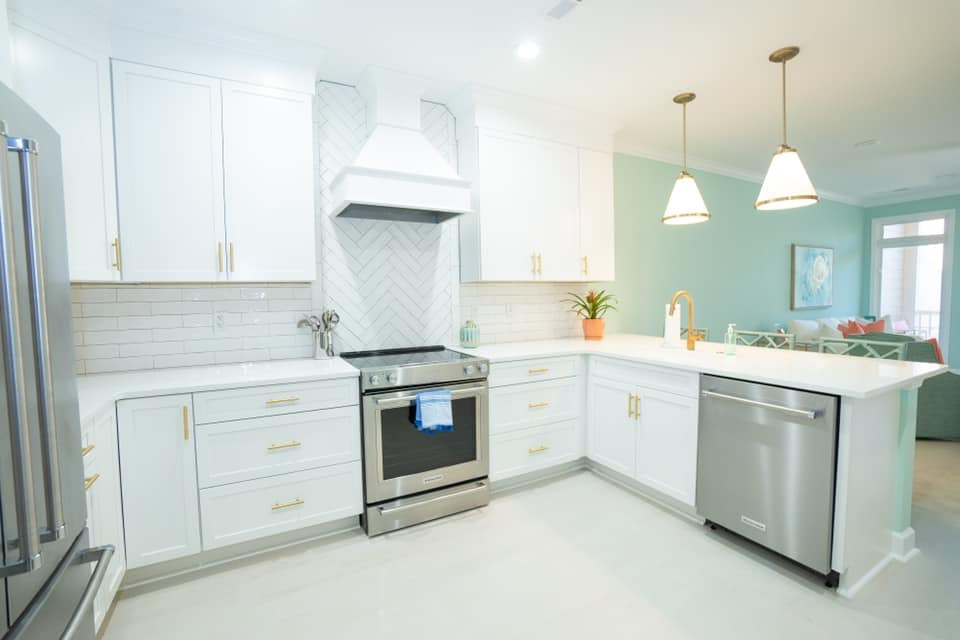 Photo of a mid-sized beach style l-shaped open plan kitchen in Charleston with a single-bowl sink, shaker cabinets, white cabinets, quartz benchtops, white splashback, ceramic splashback, stainless steel appliances, light hardwood floors, a peninsula, white floor and white benchtop.