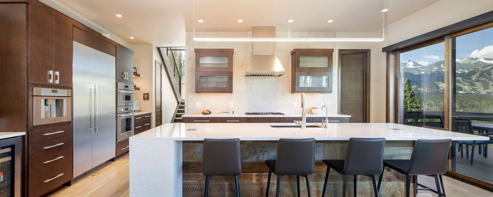 Large trendy l-shaped light wood floor open concept kitchen photo in Denver with flat-panel cabinets, medium tone wood cabinets, white backsplash, stainless steel appliances, an island and white countertops