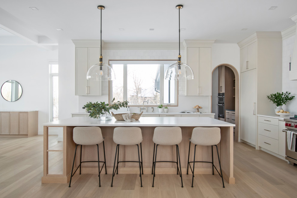 Large transitional l-shaped light wood floor kitchen photo with shaker cabinets, quartz countertops, white backsplash, quartz backsplash, stainless steel appliances, an island and white countertops