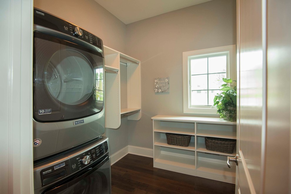 Photo of a large traditional l-shaped dedicated laundry room in Milwaukee with open cabinets, white cabinets, grey walls, dark hardwood floors, a stacked washer and dryer, brown floor, wood benchtops and white benchtop.