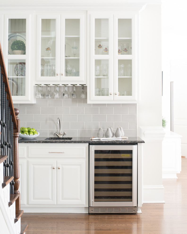 Design ideas for a mid-sized traditional single-wall wet bar in Charlotte with an undermount sink, raised-panel cabinets, white cabinets, granite benchtops, grey splashback, subway tile splashback, medium hardwood floors, brown floor and black benchtop.