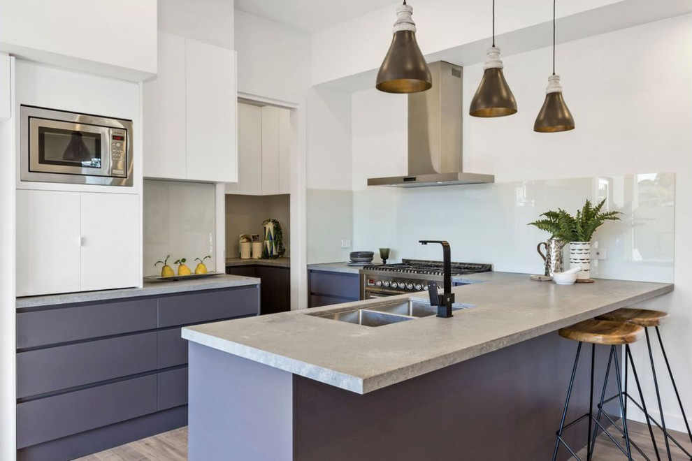 Photo of a contemporary kitchen in Melbourne with a double-bowl sink, flat-panel cabinets, white cabinets, concrete benchtops, white splashback, glass sheet splashback, stainless steel appliances, light hardwood floors, a peninsula and grey benchtop.