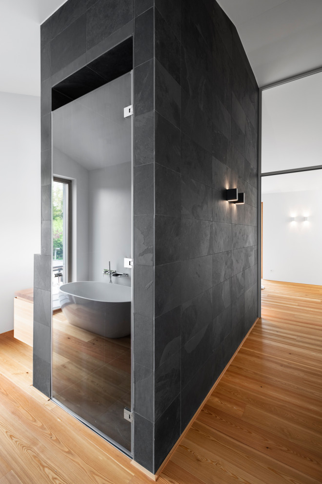 Badezimmer En Suite With flächenbündigen Schrankfronten, hellen Holzschränken, freistehender Badewanne, bodengleicher Dusche, Wandtoilette, schwarzen Fliesen, Schieferfliesen, weißer Wandfarbe, Schieferboden, Aufsatzwaschbecken, Waschtisch aus Holz, schwarzem Boden, Falttür-Duschabtrennung and brauner Waschtischplatte in Munich