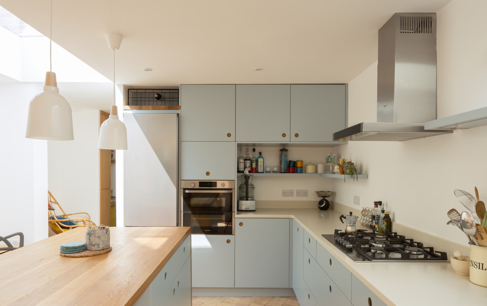 This is an example of a mid-sized contemporary l-shaped eat-in kitchen in Cambridgeshire with an integrated sink, flat-panel cabinets, blue cabinets, solid surface benchtops, white splashback, stainless steel appliances, terra-cotta floors, with island, pink floor and beige benchtop.