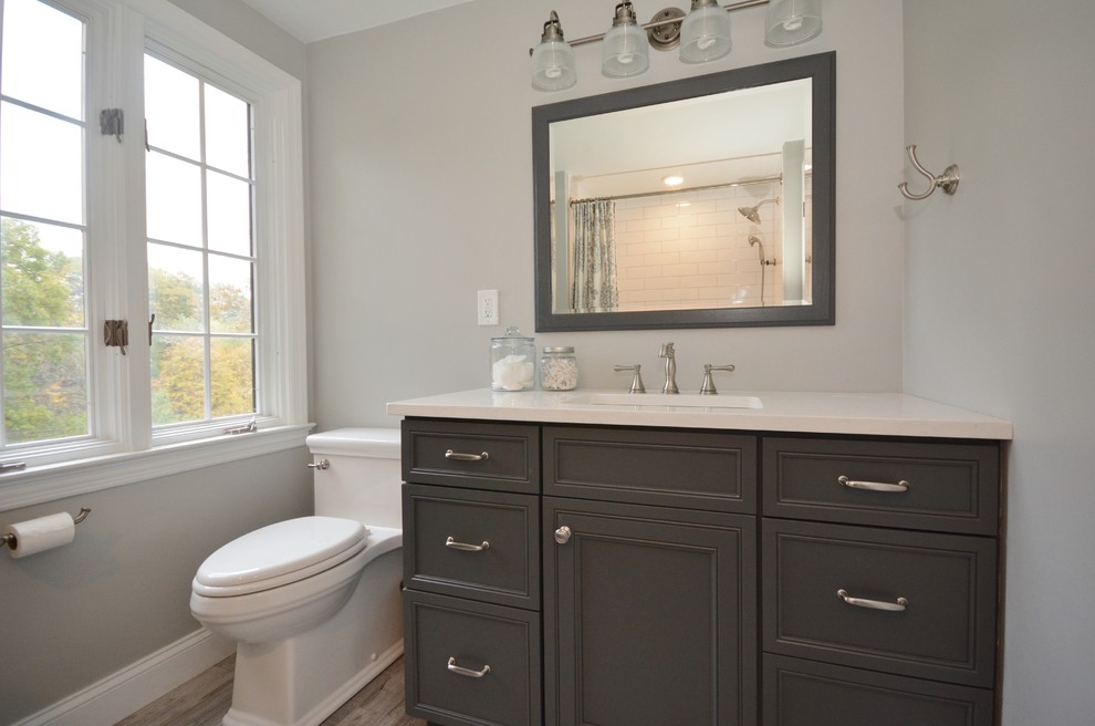 Photo of a mid-sized arts and crafts bathroom in Other with beaded inset cabinets, grey cabinets, an alcove tub, a shower/bathtub combo, a two-piece toilet, white tile, subway tile, grey walls, porcelain floors, an undermount sink, engineered quartz benchtops, beige floor, a shower curtain and white benchtops.