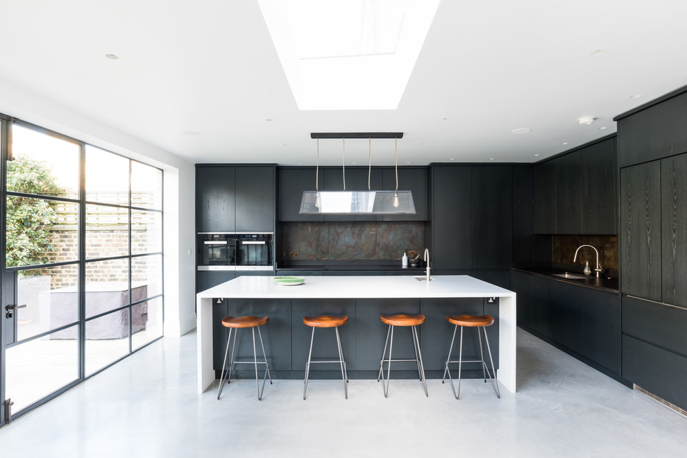 This is an example of an expansive contemporary l-shaped kitchen in London with flat-panel cabinets, quartzite benchtops, multi-coloured splashback, metal splashback, stainless steel appliances, concrete floors, with island, grey floor, white benchtop, an undermount sink and black cabinets.