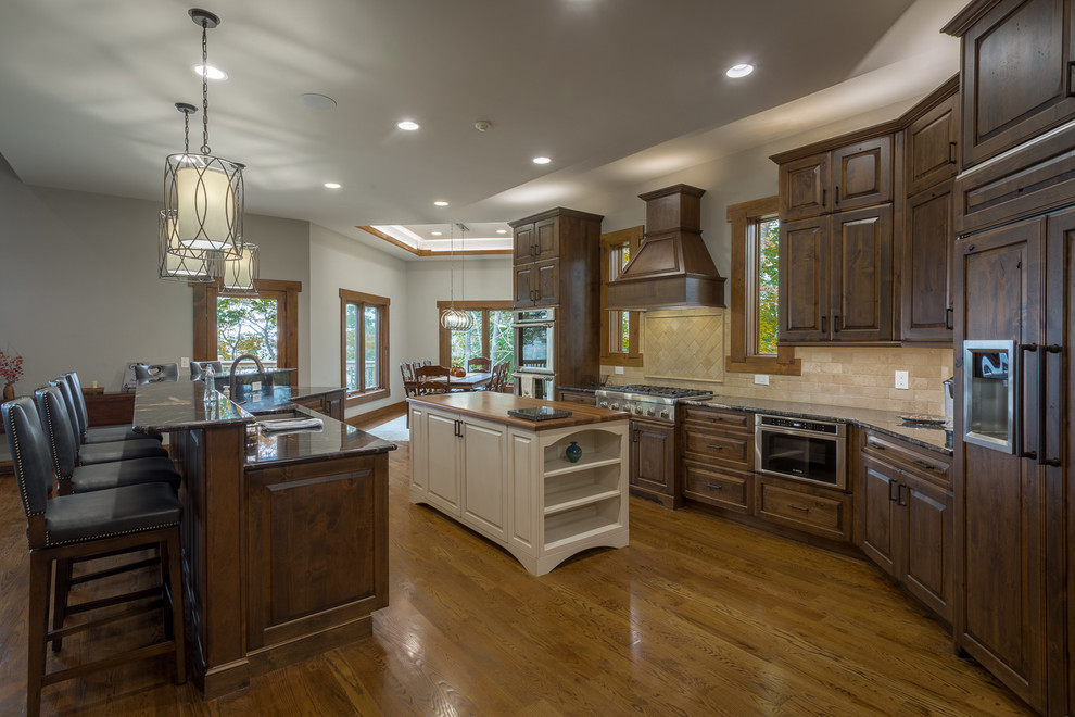 Design ideas for a large country l-shaped open plan kitchen in Charlotte with raised-panel cabinets, dark wood cabinets, granite benchtops, stone slab splashback, stainless steel appliances, medium hardwood floors, multiple islands, a double-bowl sink, beige splashback and brown floor.