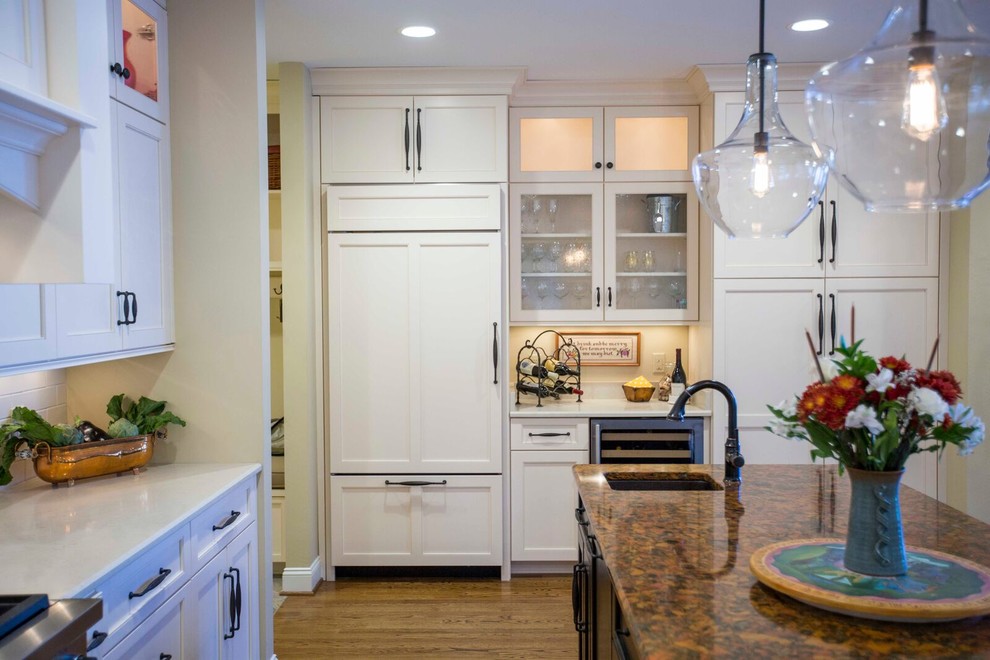 Example of a large transitional l-shaped light wood floor and brown floor enclosed kitchen design with a farmhouse sink, shaker cabinets, white cabinets, solid surface countertops, white backsplash, porcelain backsplash, stainless steel appliances and an island
