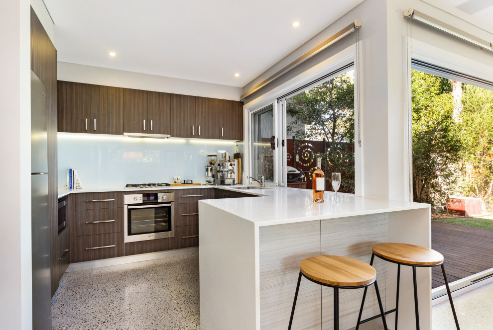 Photo of a contemporary u-shaped open plan kitchen in Sunshine Coast with brown cabinets, quartz benchtops, white splashback, porcelain splashback, stainless steel appliances, terrazzo floors, grey floor, white benchtop, an undermount sink, flat-panel cabinets and a peninsula.