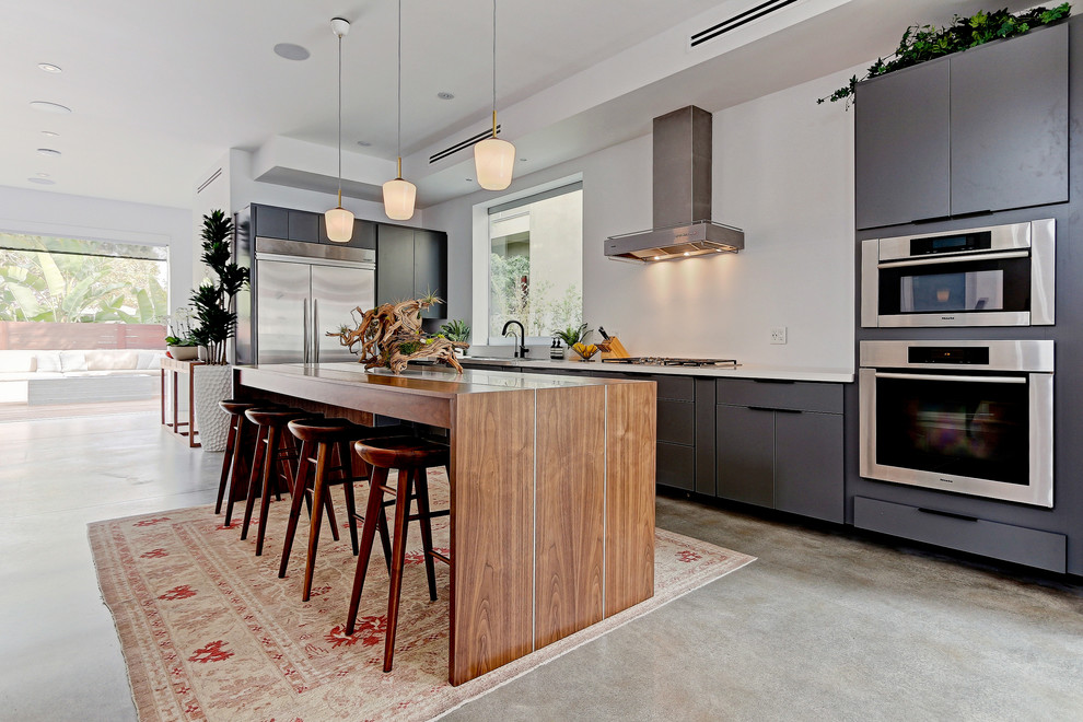 Large contemporary l-shaped open plan kitchen in Los Angeles with a drop-in sink, flat-panel cabinets, grey cabinets, solid surface benchtops, stainless steel appliances, concrete floors and with island.