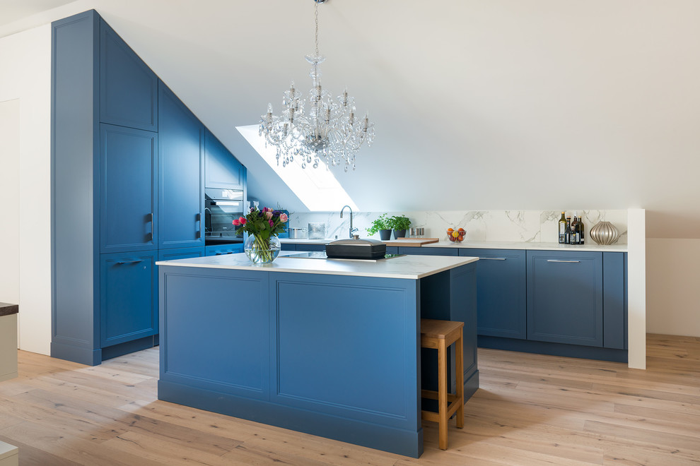 This is an example of a mid-sized transitional l-shaped kitchen in Munich with shaker cabinets, blue cabinets, white splashback, black appliances, with island, white benchtop, stone slab splashback, light hardwood floors and beige floor.