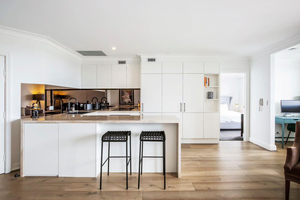 This is an example of a small contemporary u-shaped open plan kitchen in Sydney with a drop-in sink, flat-panel cabinets, white cabinets, metallic splashback, mirror splashback, black appliances, light hardwood floors, a peninsula and beige benchtop.