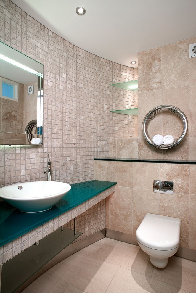 Small contemporary powder room in Dorset with open cabinets, a wall-mount toilet, beige tile, beige walls, a vessel sink, beige floor, glass benchtops and turquoise benchtops.