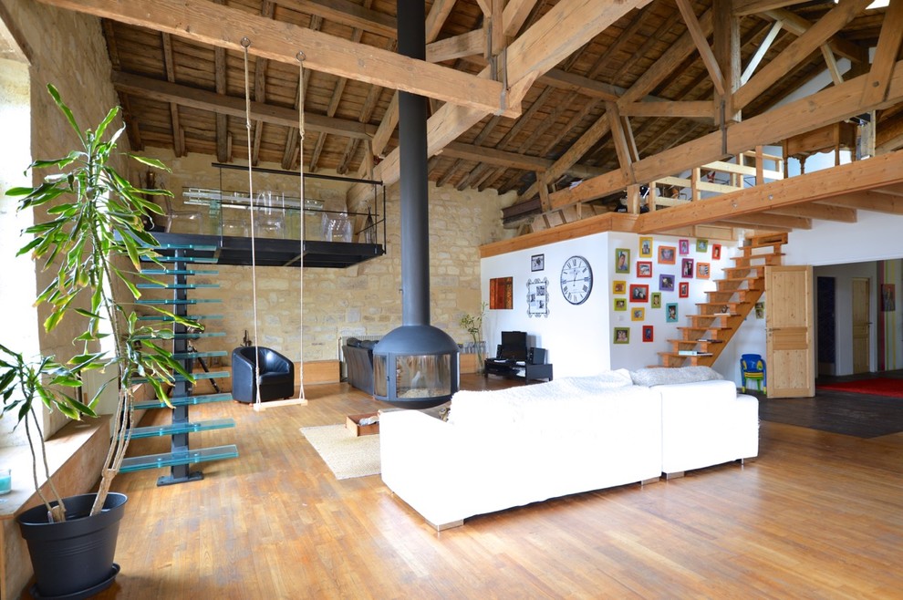 Industrial open concept family room in Bordeaux with medium hardwood floors, a hanging fireplace and white walls.