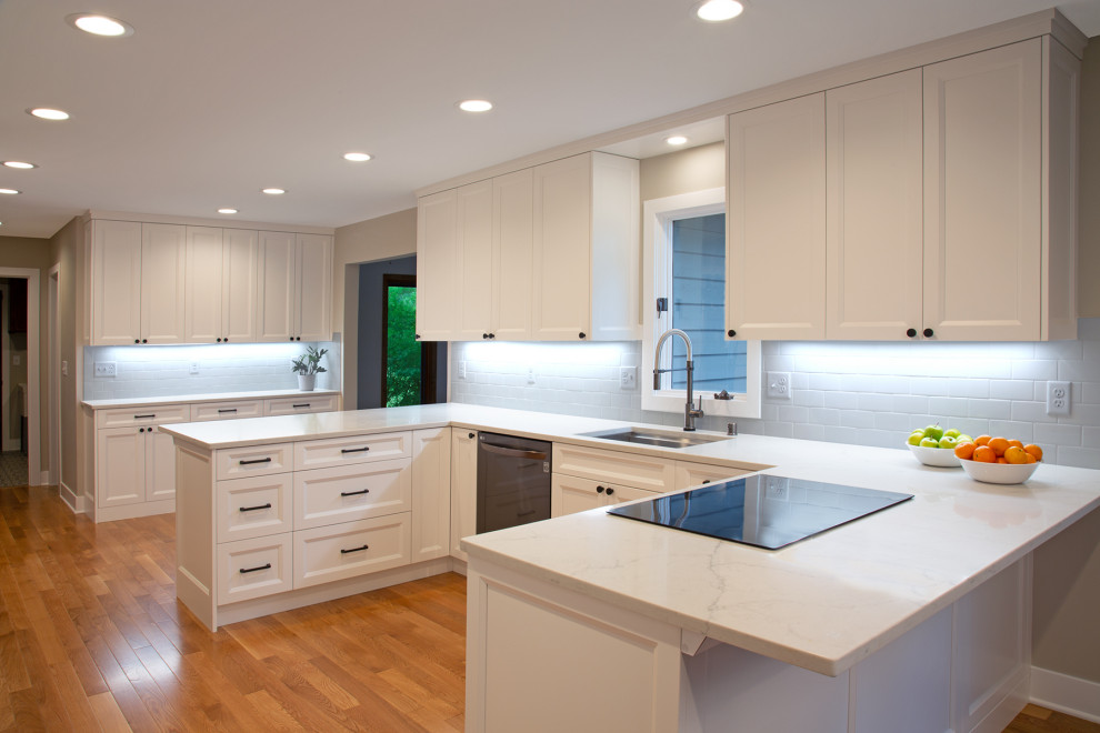 Inspiration for a transitional u-shaped eat-in kitchen in Minneapolis with recessed-panel cabinets, white cabinets, quartz benchtops, white splashback, no island, white benchtop, an undermount sink, ceramic splashback, black appliances, medium hardwood floors and brown floor.