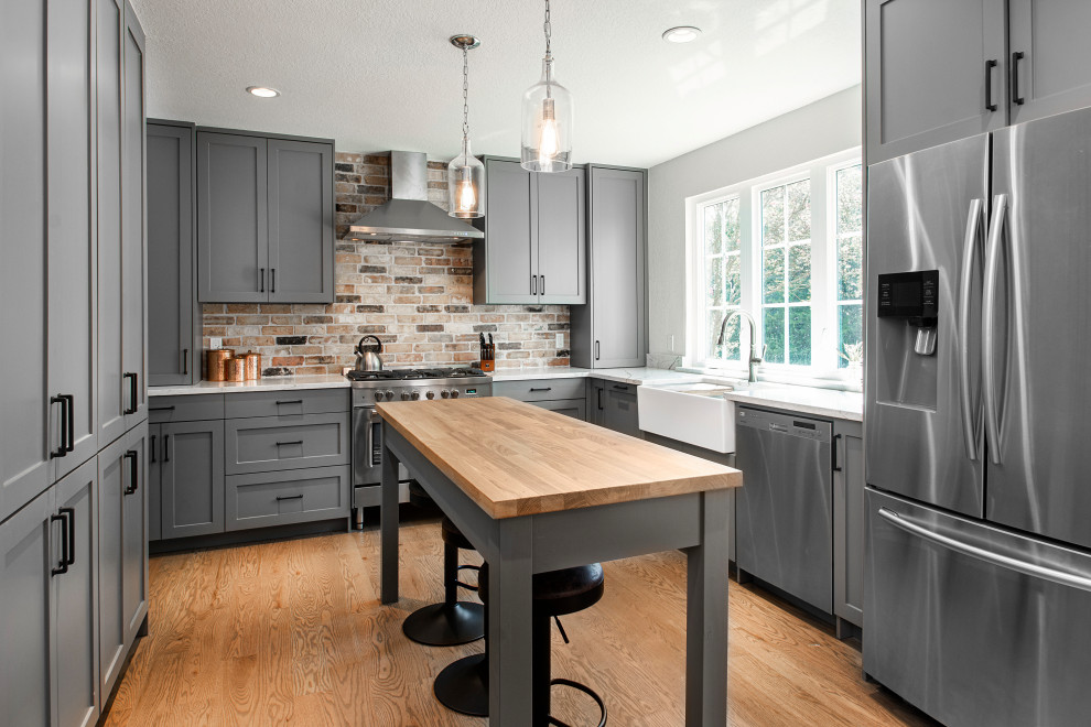 Transitional u-shaped medium tone wood floor and brown floor enclosed kitchen photo in Dallas with a farmhouse sink, shaker cabinets, gray cabinets, wood countertops, multicolored backsplash, brick backsplash, paneled appliances, an island and brown countertops