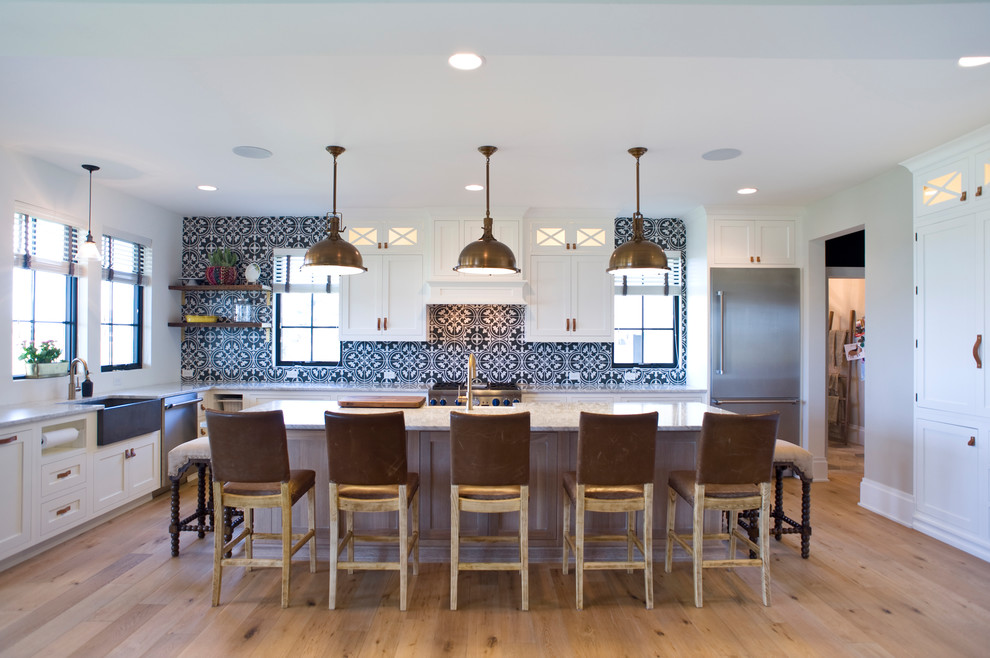 Large transitional u-shaped light wood floor and brown floor eat-in kitchen photo with a farmhouse sink, raised-panel cabinets, white cabinets, marble countertops, multicolored backsplash, porcelain backsplash, stainless steel appliances and an island