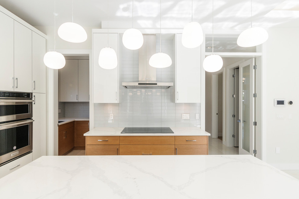This is an example of a mid-sized transitional open plan kitchen in Vancouver with an undermount sink, flat-panel cabinets, white cabinets, quartz benchtops, white splashback, glass tile splashback, stainless steel appliances, porcelain floors, with island, white floor and white benchtop.
