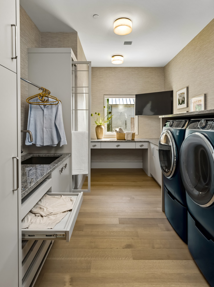 Example of a trendy u-shaped light wood floor, beige floor and wallpaper dedicated laundry room design in Philadelphia with an undermount sink, flat-panel cabinets, white cabinets, beige walls, a side-by-side washer/dryer and gray countertops