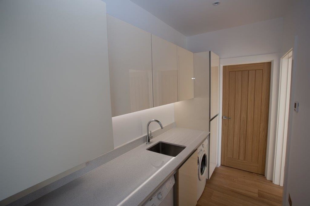 Mid-sized contemporary single-wall utility room in Essex with a farmhouse sink, flat-panel cabinets, white cabinets, solid surface benchtops, white walls, porcelain floors, a side-by-side washer and dryer, beige floor and grey benchtop.