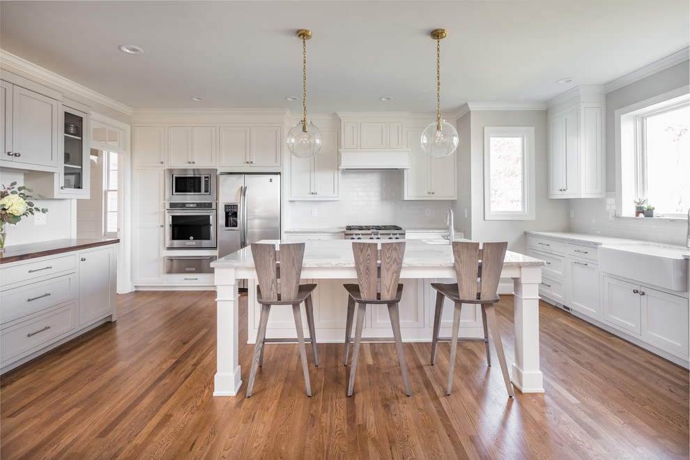 Large transitional u-shaped eat-in kitchen in Raleigh with a farmhouse sink, shaker cabinets, white cabinets, white splashback, stainless steel appliances, medium hardwood floors, with island, brown floor, white benchtop, quartzite benchtops and subway tile splashback.