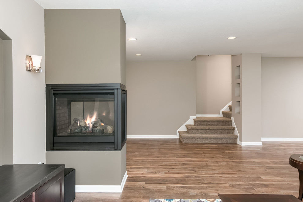 Photo of a mid-sized transitional look-out basement in Minneapolis with beige walls, medium hardwood floors, a two-sided fireplace, a plaster fireplace surround and brown floor.