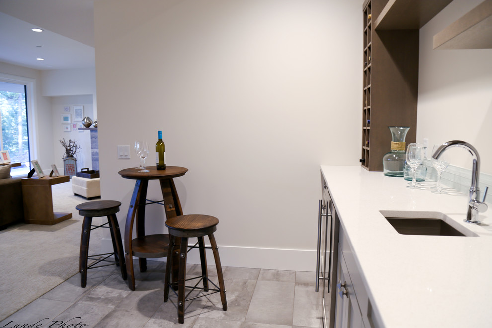 Photo of a large contemporary single-wall wet bar in Vancouver with an undermount sink, recessed-panel cabinets, beige cabinets, solid surface benchtops, white splashback, glass tile splashback and ceramic floors.