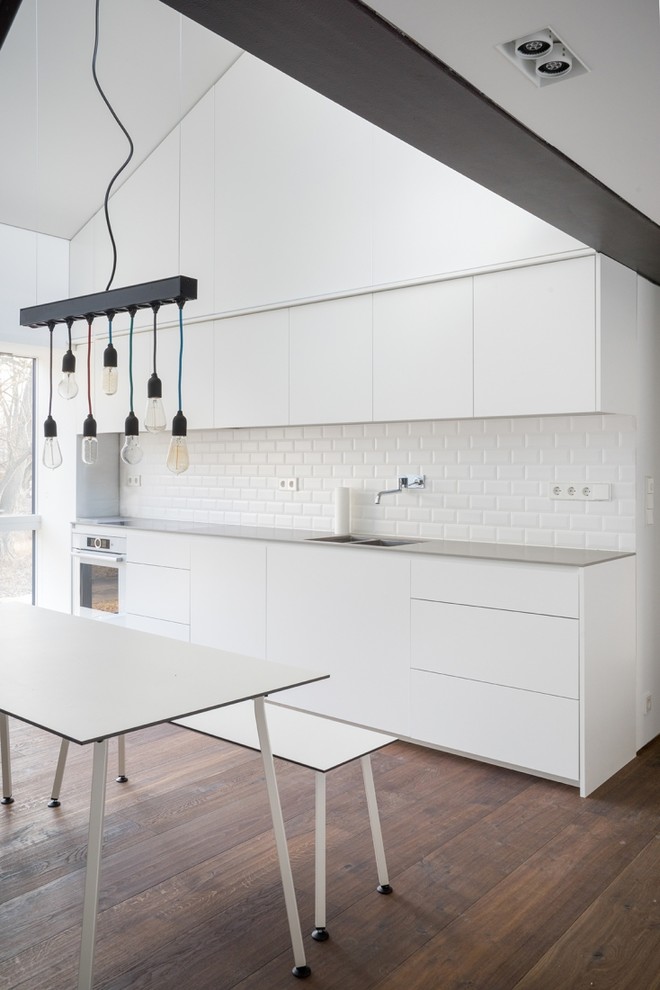 Design ideas for a mid-sized contemporary single-wall separate kitchen in Berlin with a drop-in sink, flat-panel cabinets, white cabinets, white splashback, subway tile splashback, white appliances, medium hardwood floors, no island and brown floor.