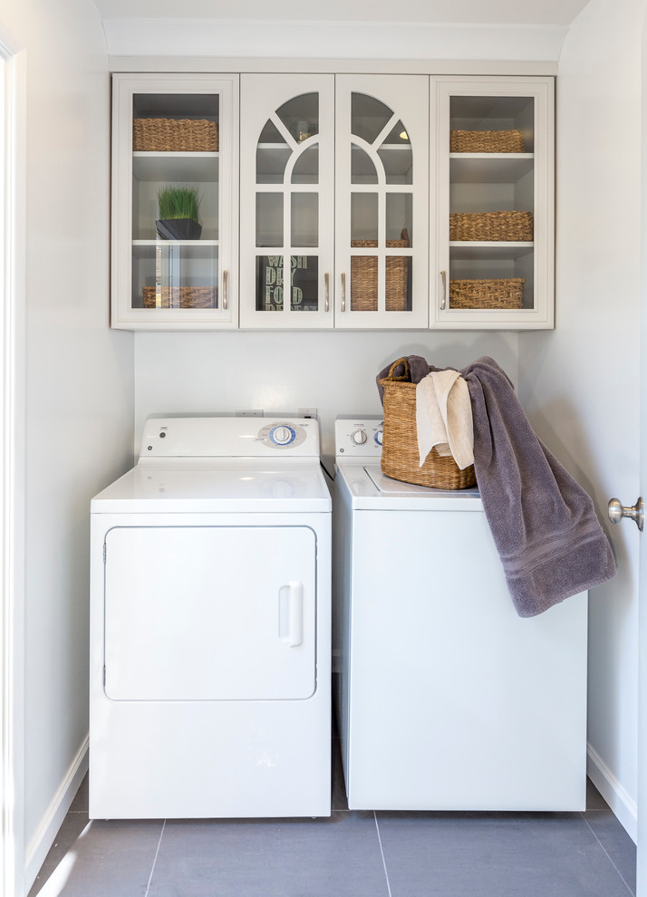 This is an example of a small transitional single-wall dedicated laundry room in Los Angeles with glass-front cabinets, white cabinets, white walls, porcelain floors, a side-by-side washer and dryer and grey floor.
