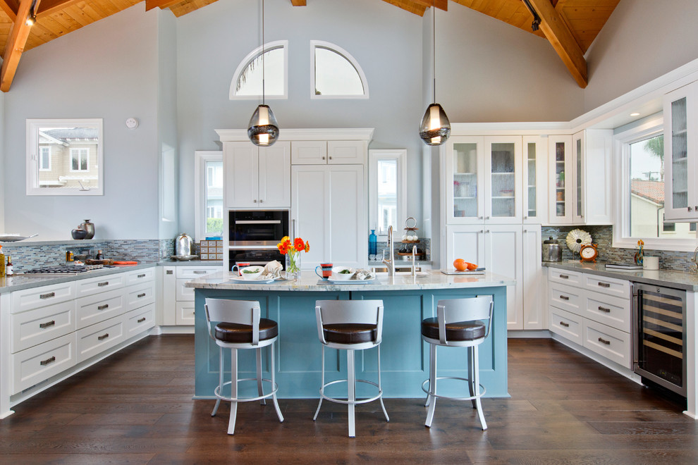 Inspiration for a large beach style u-shaped open plan kitchen in San Diego with a farmhouse sink, recessed-panel cabinets, white cabinets, quartzite benchtops, glass tile splashback, stainless steel appliances, dark hardwood floors, with island and multi-coloured splashback.
