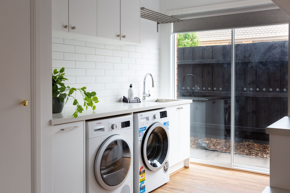 Photo of a medium sized modern galley utility room in Melbourne with a built-in sink, shaker cabinets, white cabinets, composite countertops, white splashback, metro tiled splashback, light hardwood flooring, a side by side washer and dryer and white worktops.