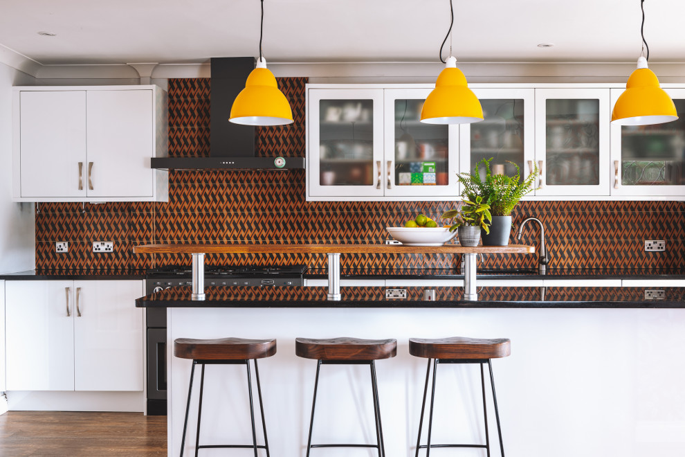 Photo of a mid-sized contemporary single-wall eat-in kitchen in Berkshire with an undermount sink, flat-panel cabinets, white cabinets, granite benchtops, orange splashback, cement tile splashback, coloured appliances, vinyl floors, with island, brown floor and black benchtop.