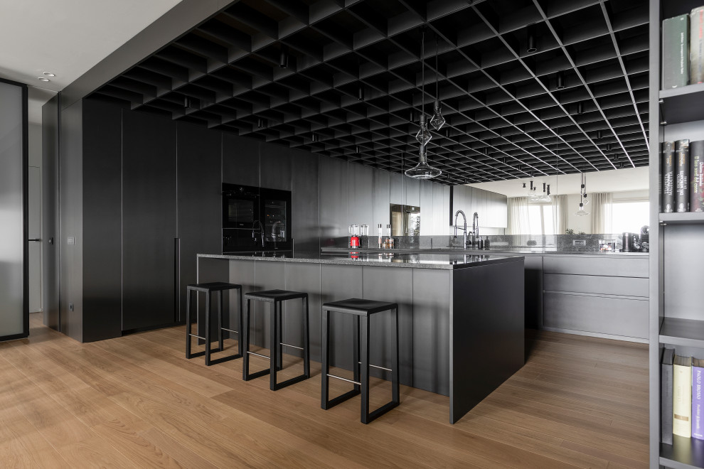 Photo of an expansive modern u-shaped open plan kitchen in Milan with a drop-in sink, flat-panel cabinets, granite benchtops, black splashback, granite splashback, black appliances, light hardwood floors, with island, black benchtop, coffered, grey cabinets and beige floor.