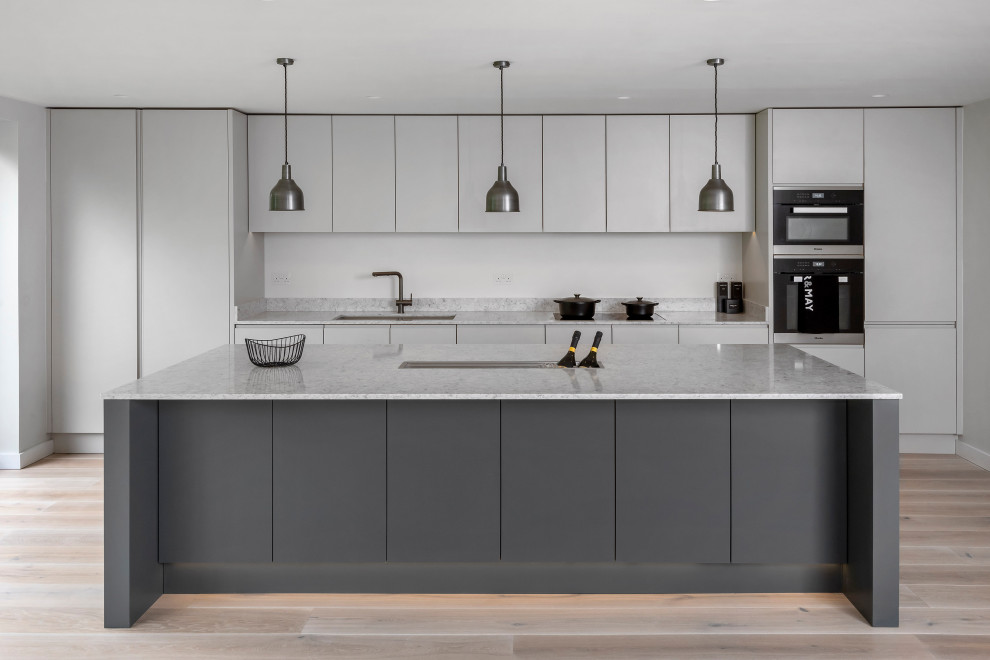 Design ideas for a large modern galley open plan kitchen with a submerged sink, flat-panel cabinets, grey cabinets, composite countertops, black appliances, light hardwood flooring, an island, beige floors and grey worktops.