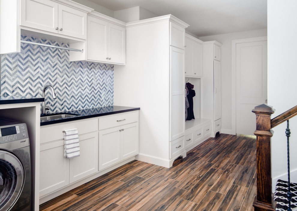 This is an example of a large transitional single-wall laundry room in Houston with shaker cabinets, white cabinets, granite benchtops, stone tile splashback, white walls, porcelain floors, a side-by-side washer and dryer, brown floor, black benchtop, multi-coloured splashback and an undermount sink.