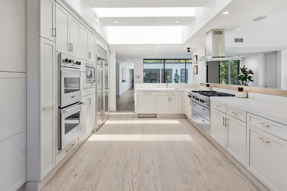 Photo of a contemporary kitchen in Los Angeles with shaker cabinets, white cabinets, white appliances, light hardwood floors and white benchtop.