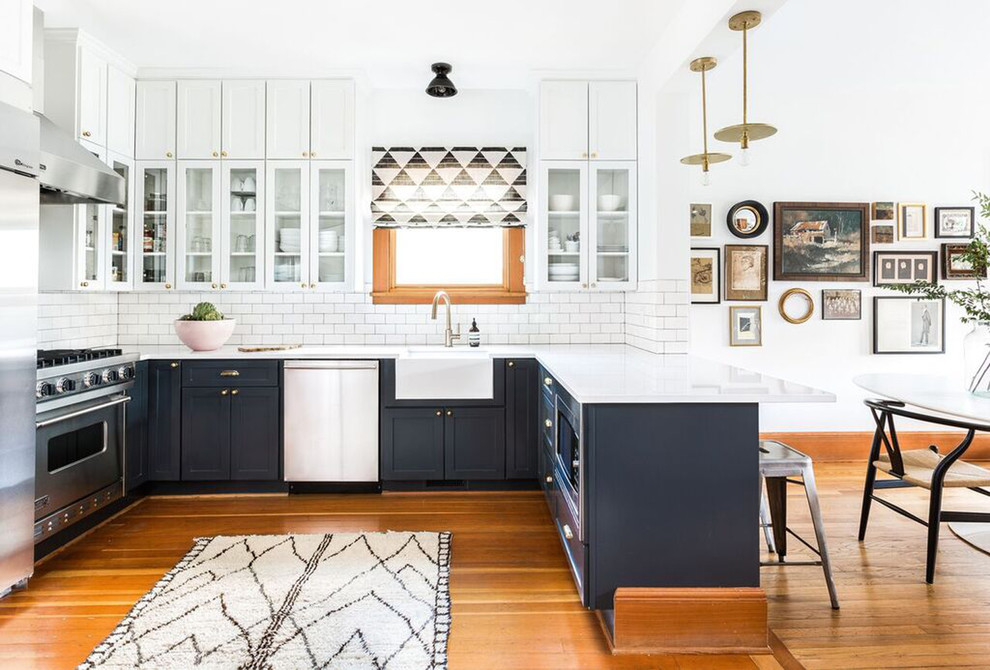 Inspiration for a mid-sized country u-shaped open plan kitchen in Seattle with a farmhouse sink, shaker cabinets, grey cabinets, white splashback, subway tile splashback, stainless steel appliances, medium hardwood floors, a peninsula, quartzite benchtops and brown floor.