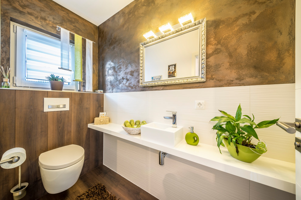 Moderne Gästetoilette mit Wandtoilette, weißen Fliesen, brauner Wandfarbe, dunklem Holzboden, Aufsatzwaschbecken, braunem Boden und weißer Waschtischplatte in Sonstige
