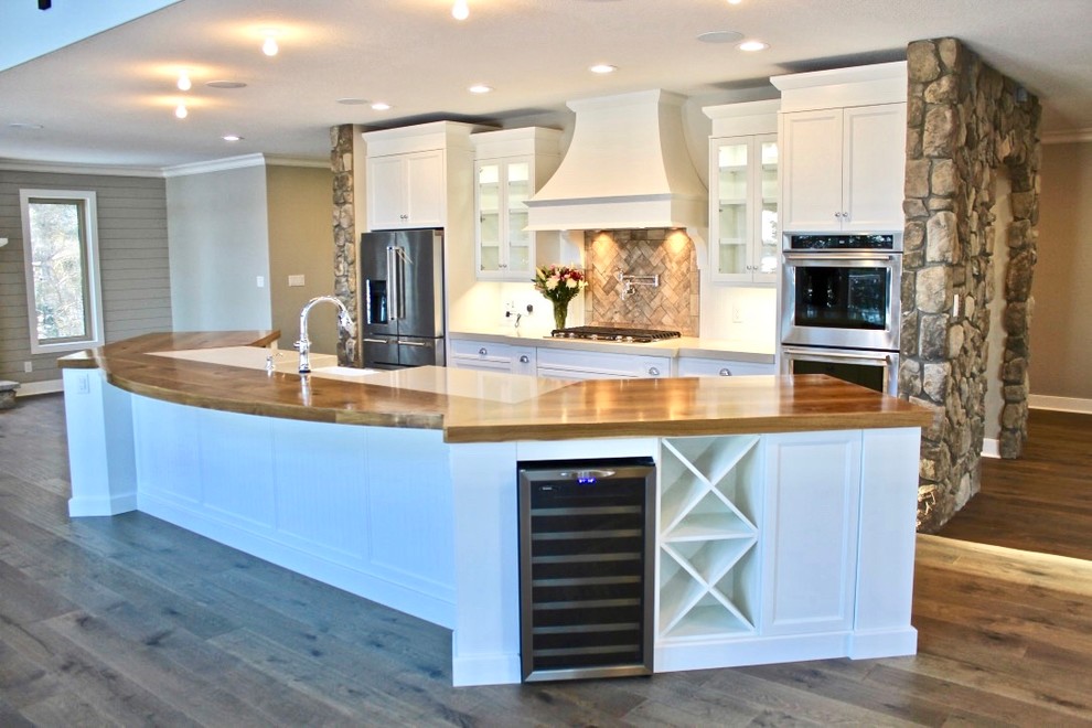 Photo of a beach style single-wall open plan kitchen in Other with a farmhouse sink, shaker cabinets, white cabinets, quartz benchtops, white splashback, timber splashback, stainless steel appliances, medium hardwood floors, with island, brown floor and beige benchtop.