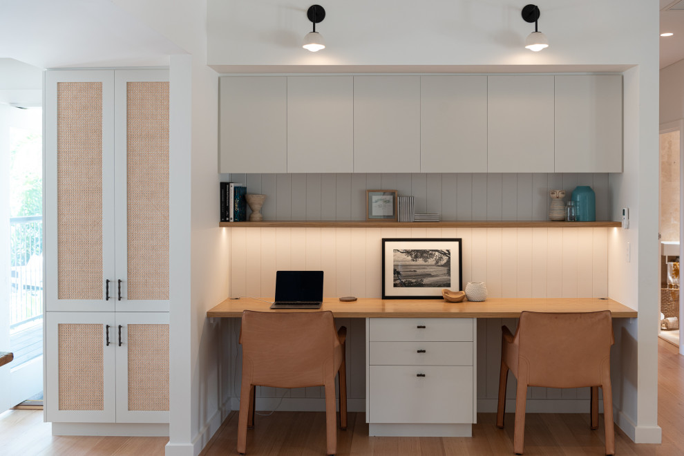 Diseño de despacho marinero sin chimenea con paredes blancas, suelo de madera en tonos medios, escritorio empotrado, suelo marrón y panelado