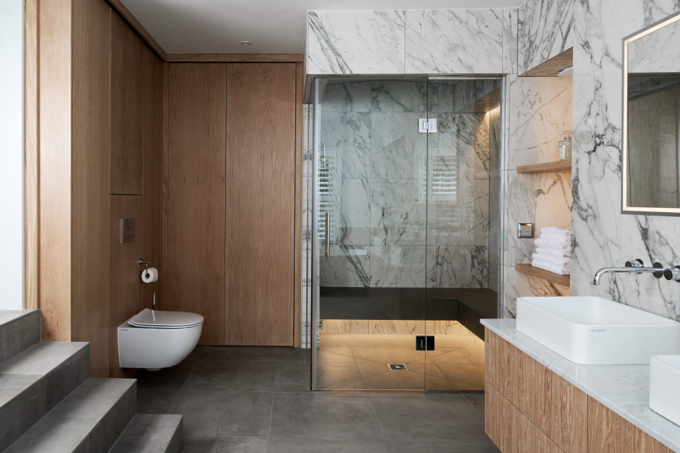 Modern inredning av ett stort vit vitt en-suite badrum, med släta luckor, skåp i ljust trä, en vägghängd toalettstol, grå kakel, marmorkakel, klinkergolv i keramik, grått golv, dusch med gångjärnsdörr, en kantlös dusch och ett fristående handfat