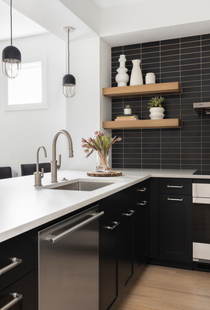 This is an example of a large modern u-shaped open plan kitchen in Portland with a submerged sink, shaker cabinets, black cabinets, engineered stone countertops, black splashback, porcelain splashback, stainless steel appliances, light hardwood flooring, a breakfast bar and white worktops.